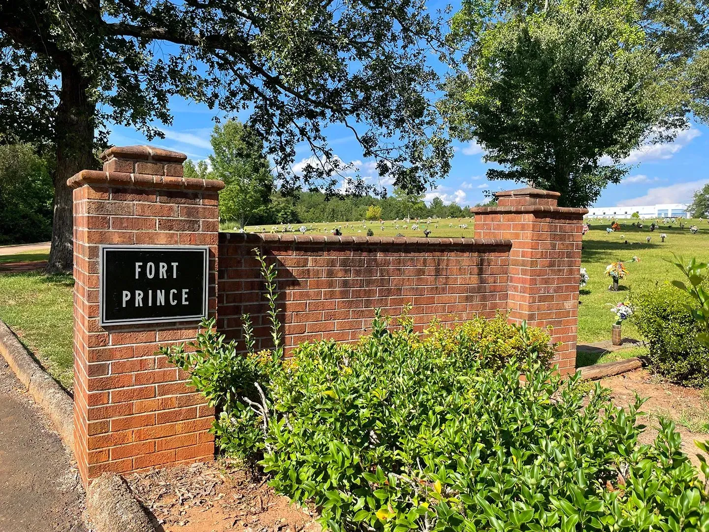 Brick Fort Prince sign entrance with brick wall, greenery, and trees under a blue sky