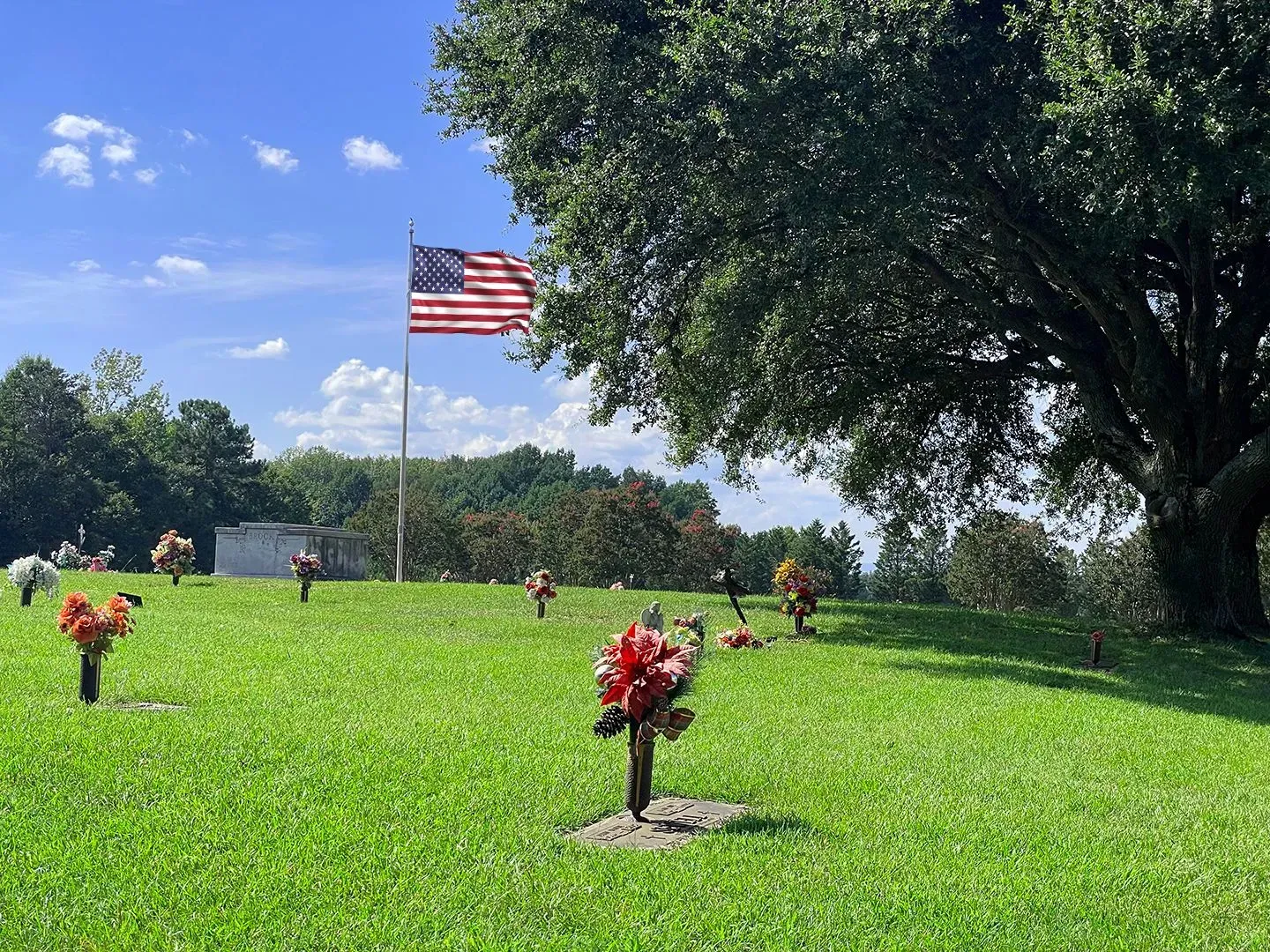 Grassy memorial with red flower markers and a U.S. flag waving under a blue sky