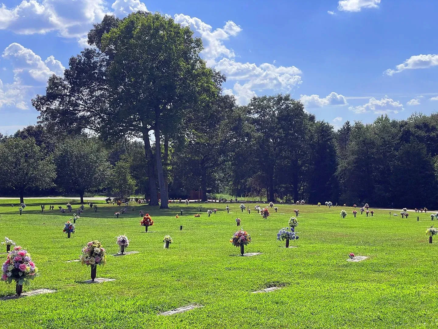 Rows of flower bouquets in a green cemetery under tall trees and a partly cloudy blue sky