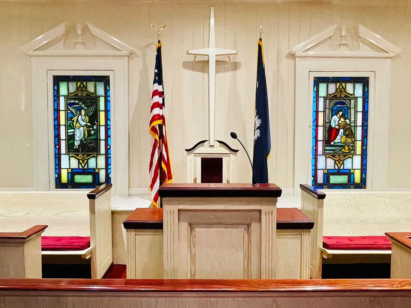 Church altar with cross, American flag, blue flag, stained-glass windows, and red-cushioned pews