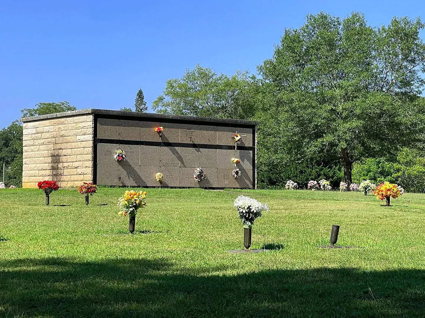 Memorial garden with small flower arrangements on a grassy lawn beside a stone wall and trees