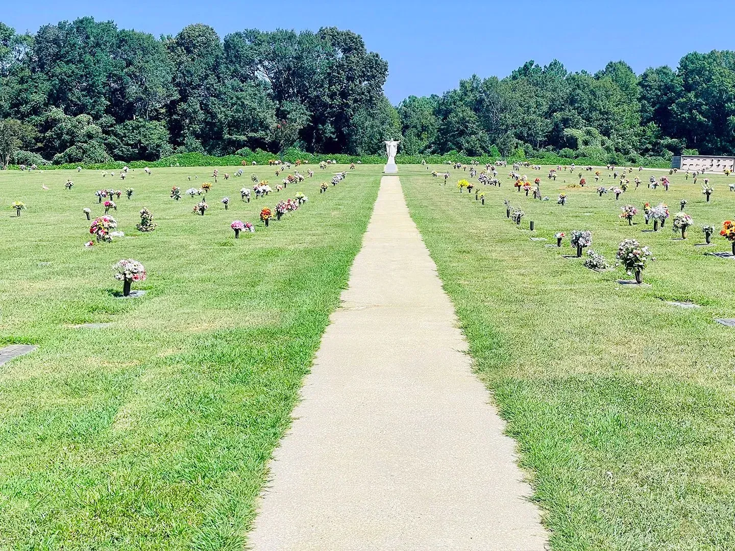A long path through a cemetery with rows of small headstones on green grass under a blue sky