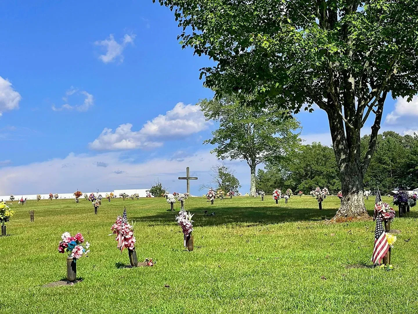 Grassy cemetery with small flower bouquets, a large tree, and a cross under a blue sky with clouds