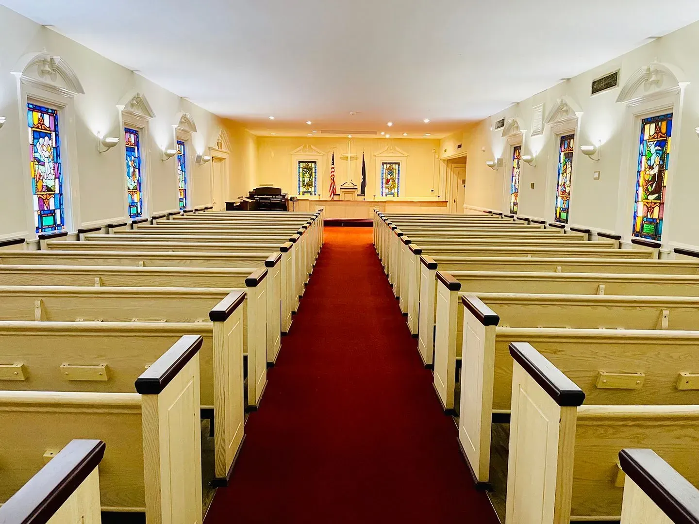 Empty church pews facing a red carpet aisle in a bright sanctuary with stained glass windows