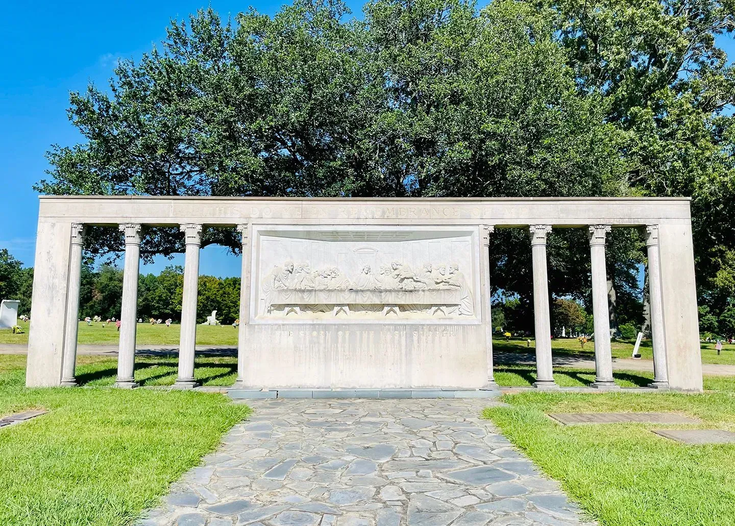 Stone memorial wall with engraved plaque, framed by white columns and trees under a blue sky