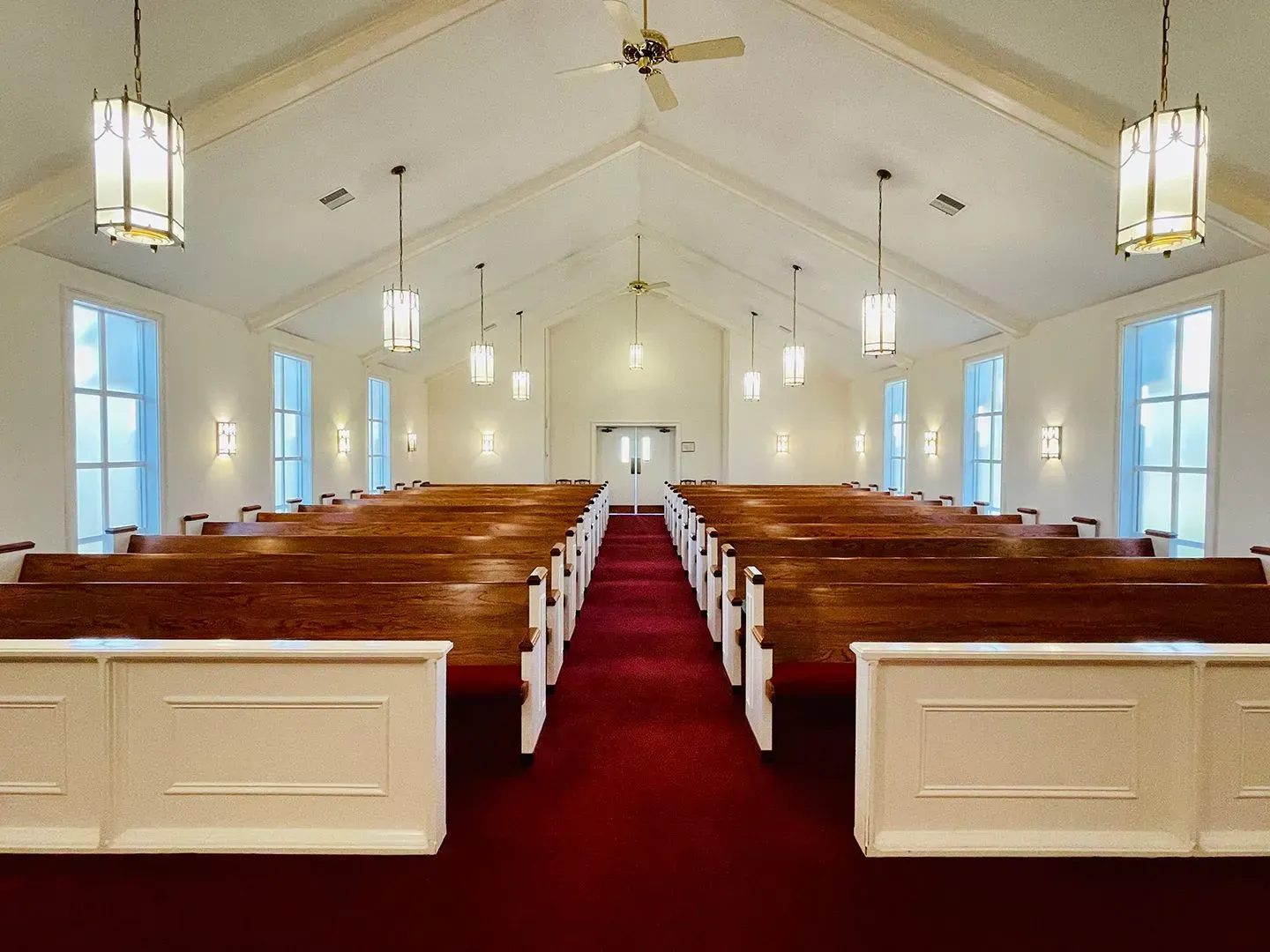 Empty white church sanctuary with red carpet aisle, wooden pews, and hanging lantern lights