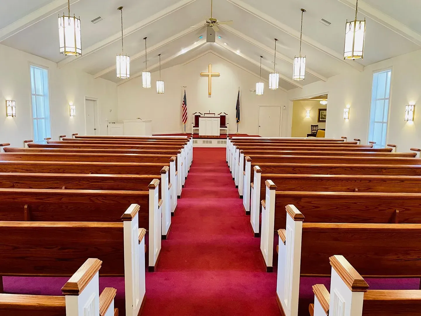 Empty church sanctuary with red carpet aisle, wooden pews, white walls, and hanging pendant lights