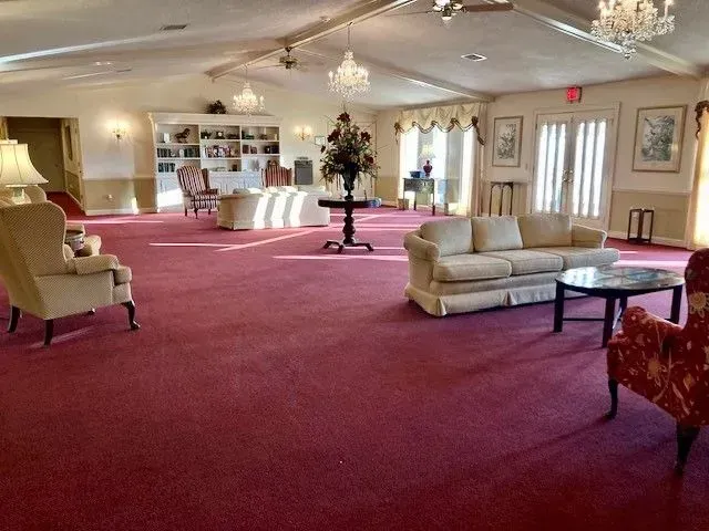 Spacious lounge with burgundy carpet, beige sofas, chairs, chandeliers, and windows with curtains