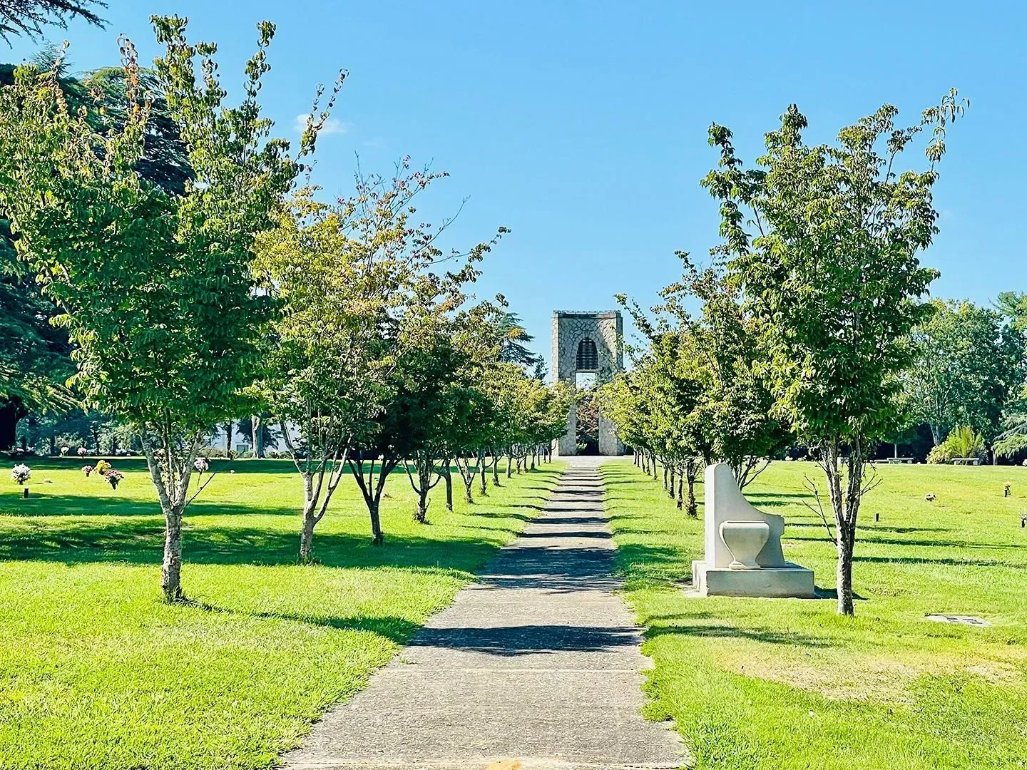 Tree-lined path through a bright green park leading to a stone monument under a blue sky