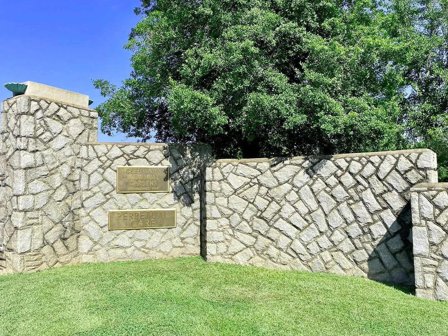 Stone wall with a gate and plaques in a grassy yard, with a large tree behind it.
