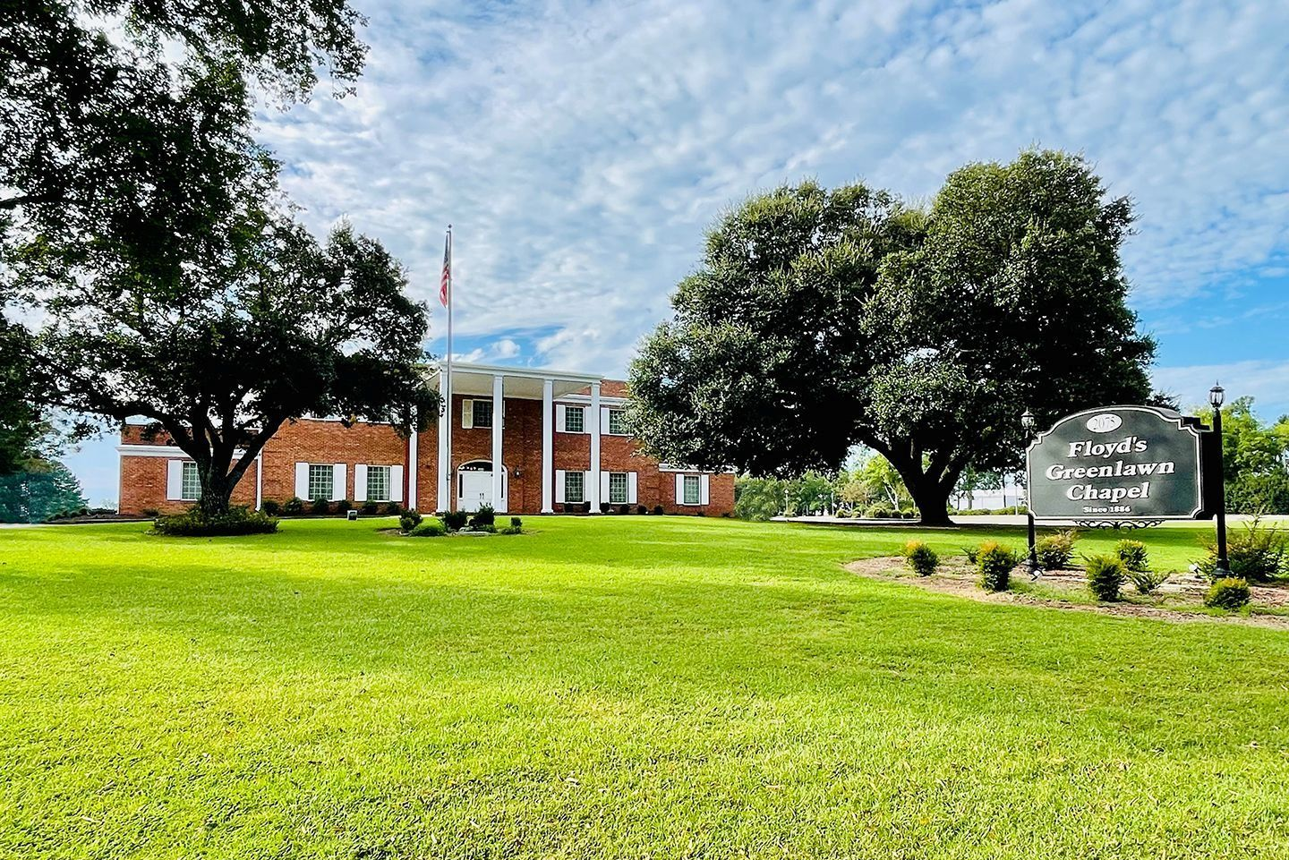Brick school building with white columns, American flag, and sign on a green lawn under trees