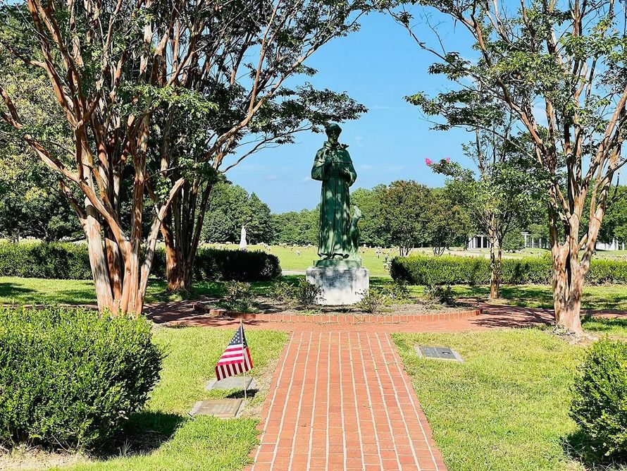 Statue at the end of a brick path in a sunny park, framed by trees and green hedges
