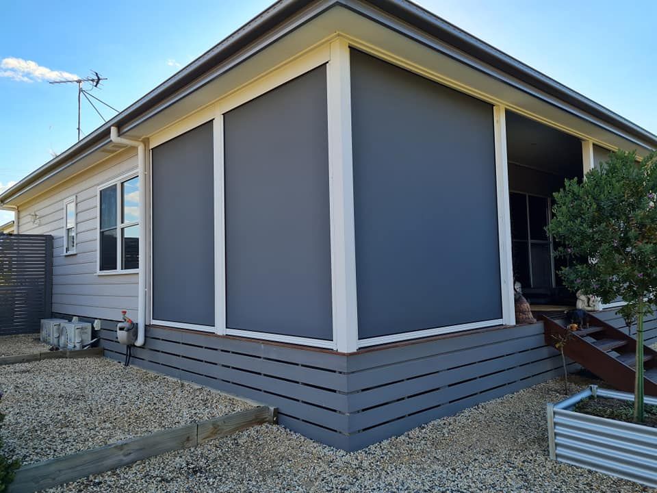 House with grey siding, corner deck with dark grey blinds, gravel, and small tree.