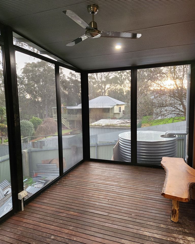 Enclosed porch with a wood deck, black-framed windows, and a tree trunk bench. The background includes houses and a water tank.
