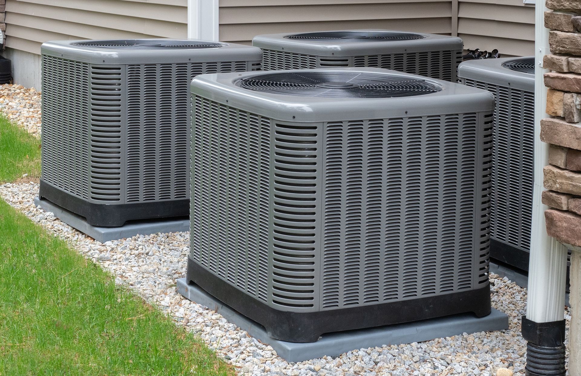 Three gray outdoor air conditioning units beside a house on a gravel bed