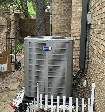Outdoor HVAC condenser unit beside a brick house, with shrubs and a white fence in front