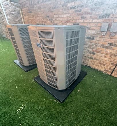 Two outdoor HVAC units beside a brick wall on artificial grass mats