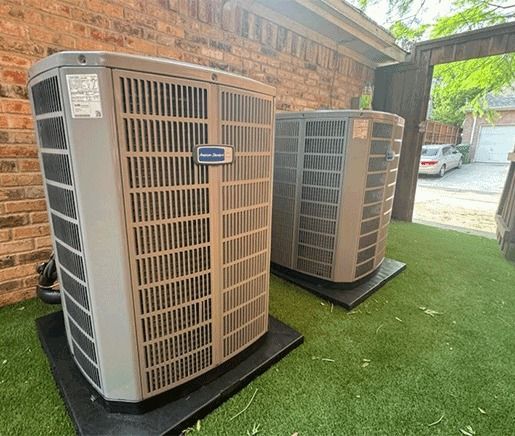 Two outdoor HVAC units on black pads beside a brick wall and green turf.
