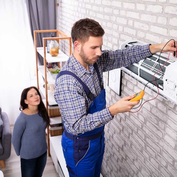 Electrician testing exposed wall wiring while a woman watches in a room with a brick wall.