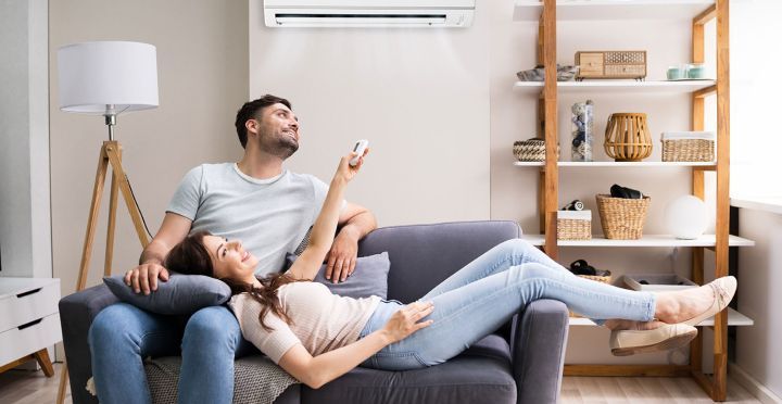 Couple relaxing on a couch in a cozy living room, with a floor lamp, shelves, and air conditioner behind them.
