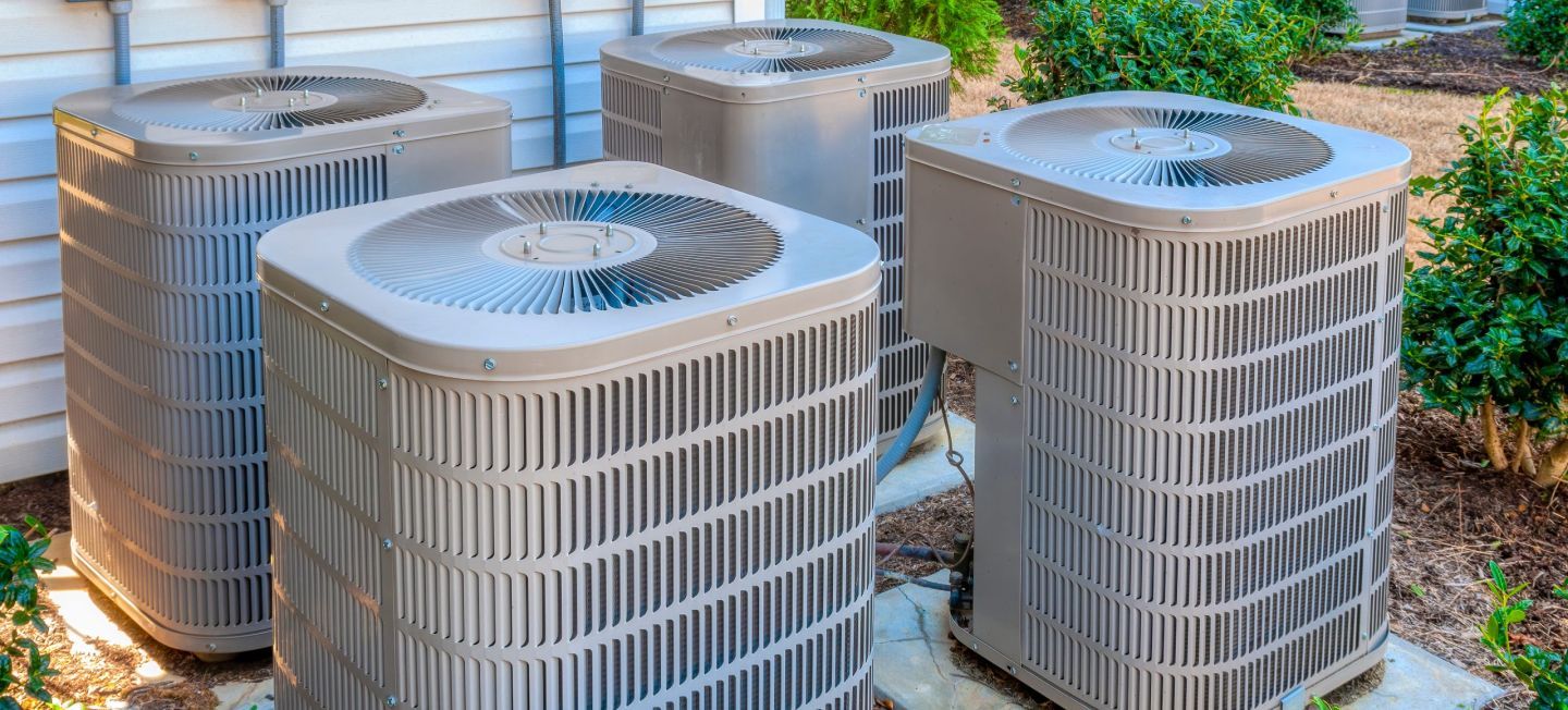 Five outdoor HVAC units on a gravel pad beside a building, with greenery in the background