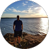 Person standing on a rocky shore facing a calm sea at sunset