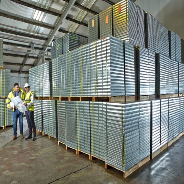Two construction professionals in hard hats review plans next to tall, stacked rows of corrugated metal roofing sheets.