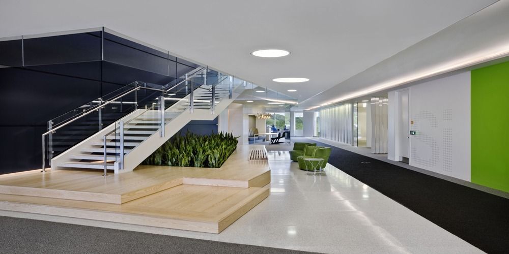 A modern office lobby features a wooden staircase, indoor plants, a white tiled floor, and green and dark blue accents.