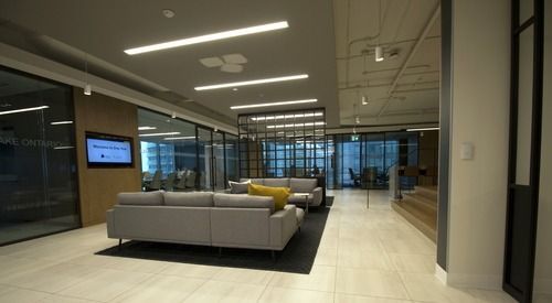 A modern office lobby with a gray sofa, a yellow throw pillow, and glass-walled conference rooms.