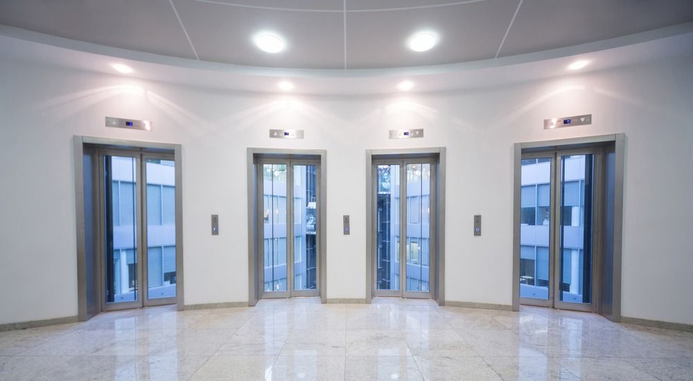 Four modern glass elevators arranged in a curved line on a white, marble-floored hall with recessed ceiling lighting.