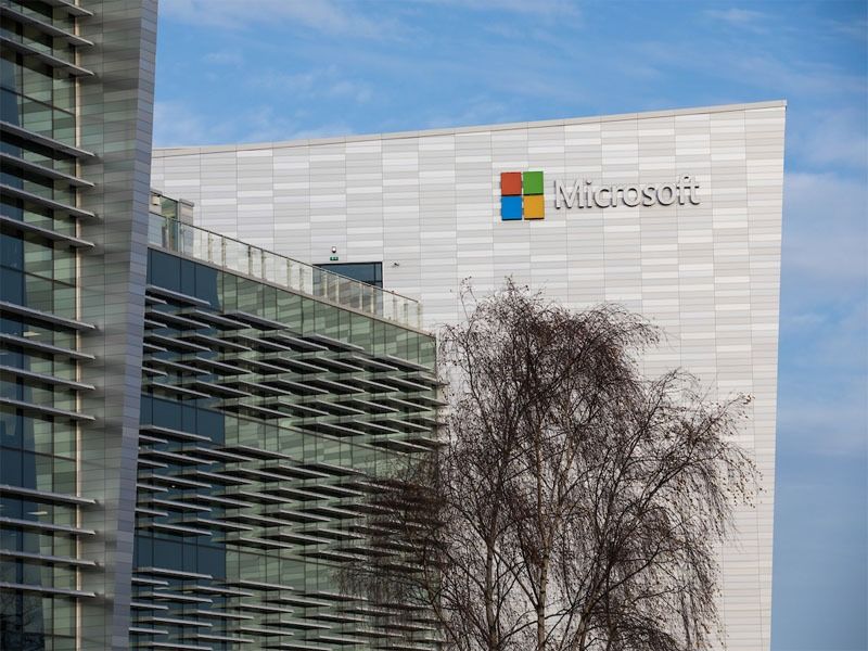 The Microsoft logo on a white modern office building facade, with glass windows and a bare tree in the foreground.