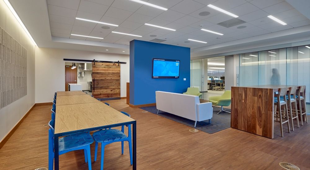 A modern office lounge with a wooden communal table, blue chairs, a blue wall with a television, and a white sofa.