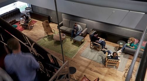 An elevated view of a modern lounge with people relaxing on sofas and chairs across several rugs on a wooden floor.