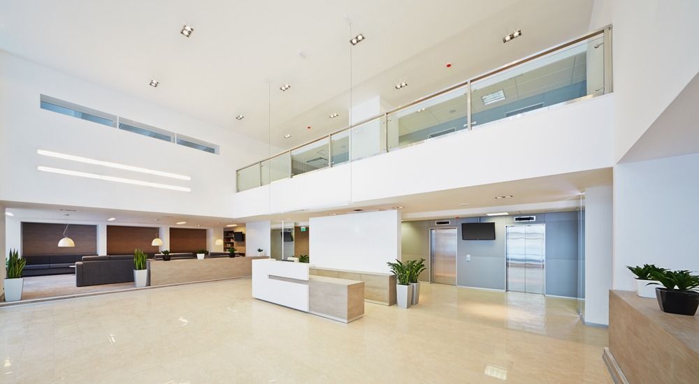 A bright, modern office lobby with a central reception desk, light-colored tiled floors, and a glass-railed mezzanine.