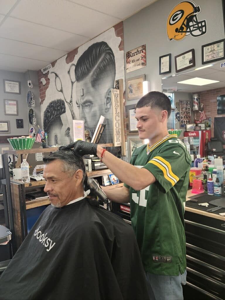 A man is getting his hair cut by a barber wearing a green bay packers jersey.