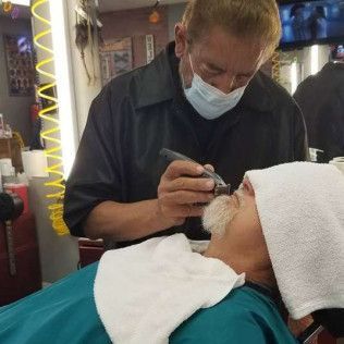 A man wearing a mask is shaving another man 's face in a barber shop.