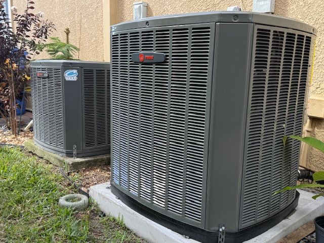 Two outdoor air conditioning units on concrete pads next to a building with brown siding.