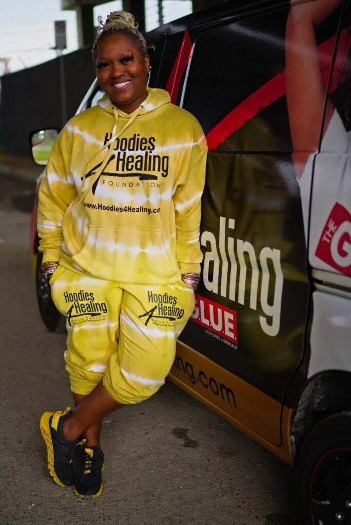 A woman in a yellow tie dye hoodie is standing next to a van.