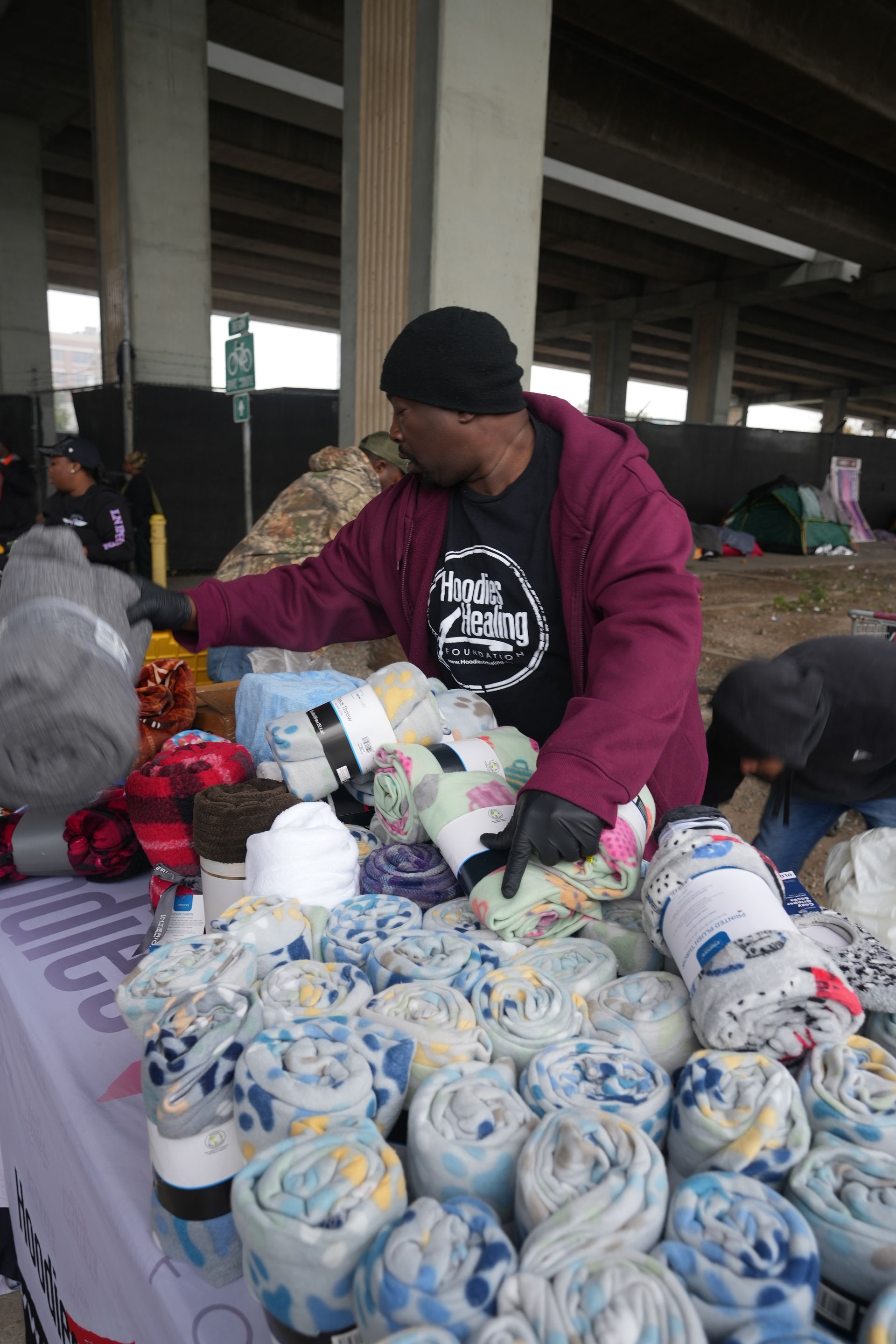 A man in a purple jacket is standing in front of a table full of blankets.