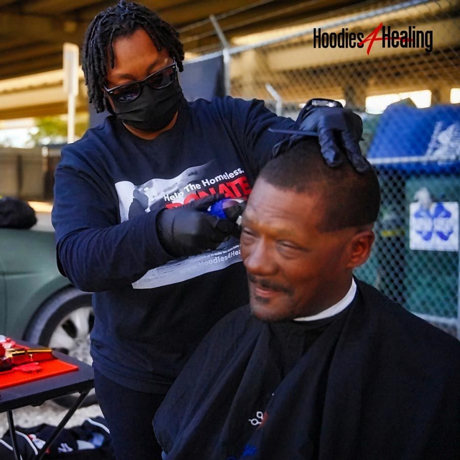 A man is getting his hair cut by a man wearing a shirt that says hoodies healing