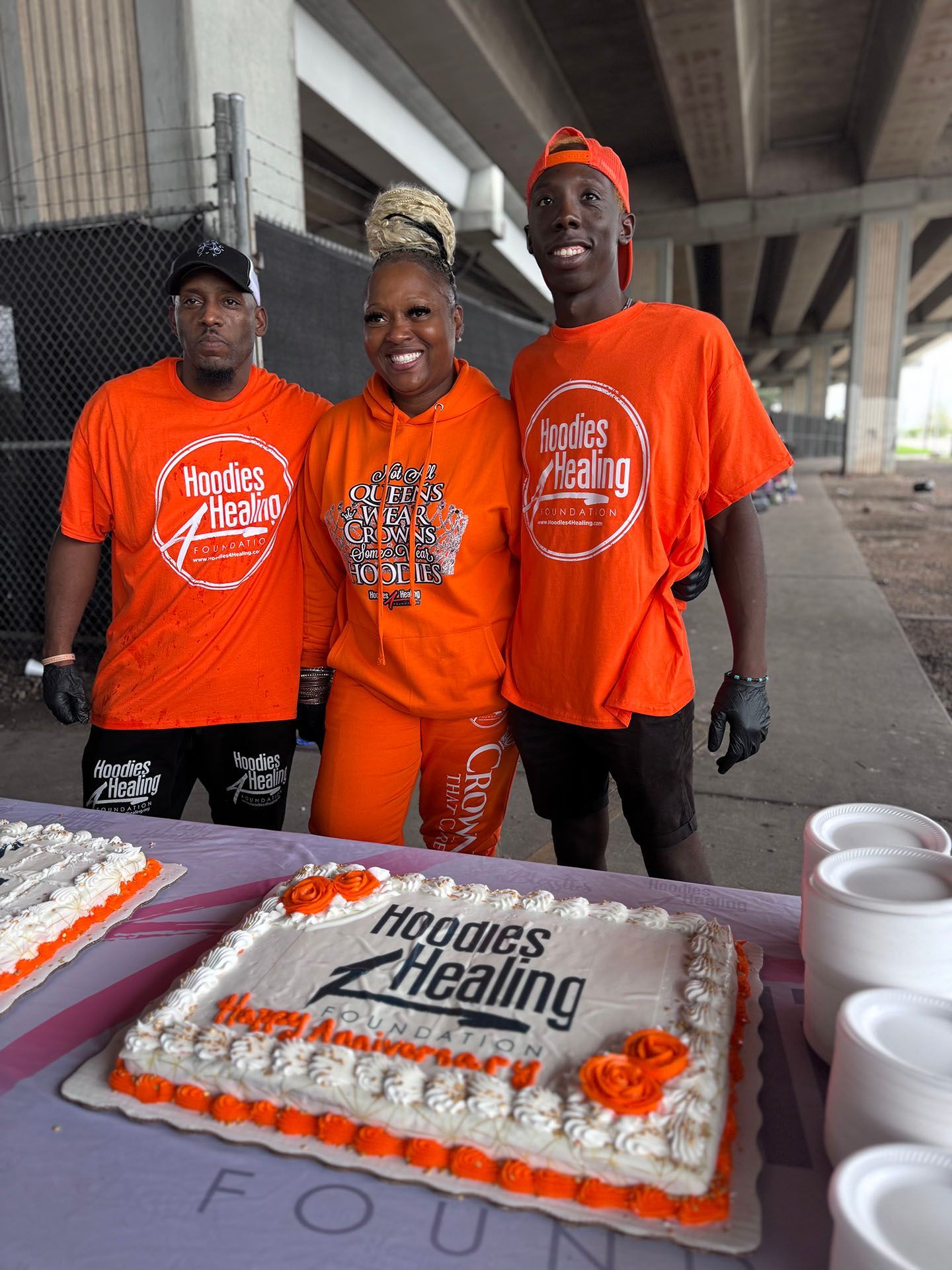 Three people in orange shirts are standing next to a cake on a table.