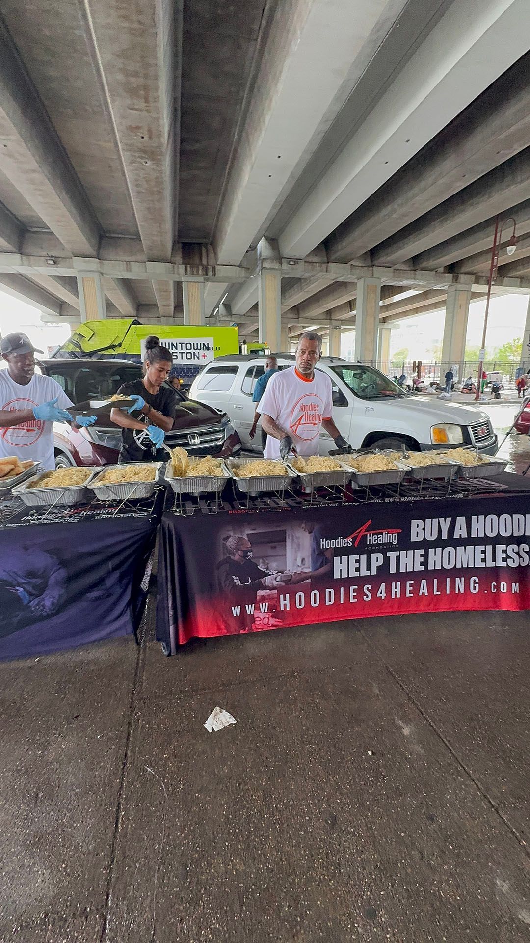 A group of people are standing around a table serving food under a bridge.