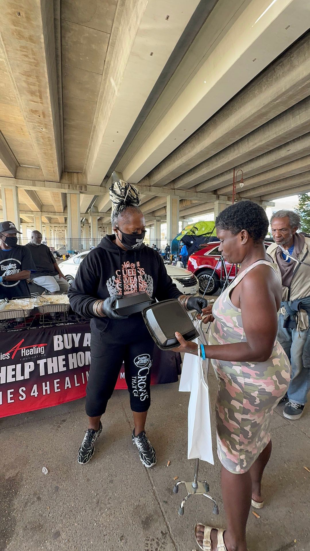 A woman is giving a box of food to another woman under a bridge.