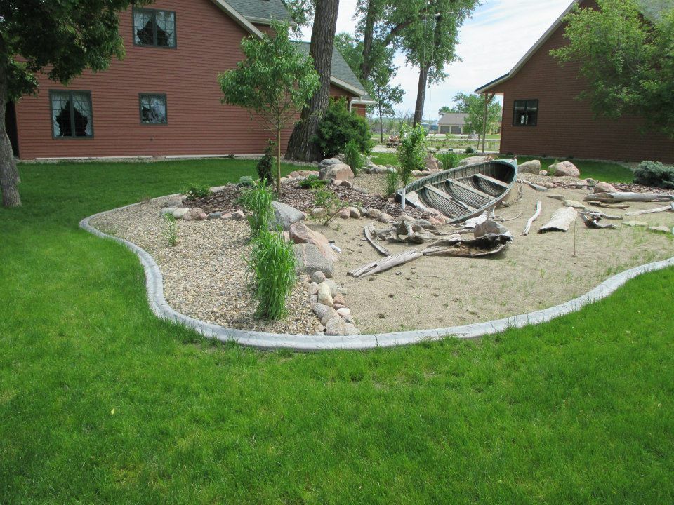 A garden bed with a decorative weathered boat surrounded by sand and greenery, set between brown buildings and green grass.