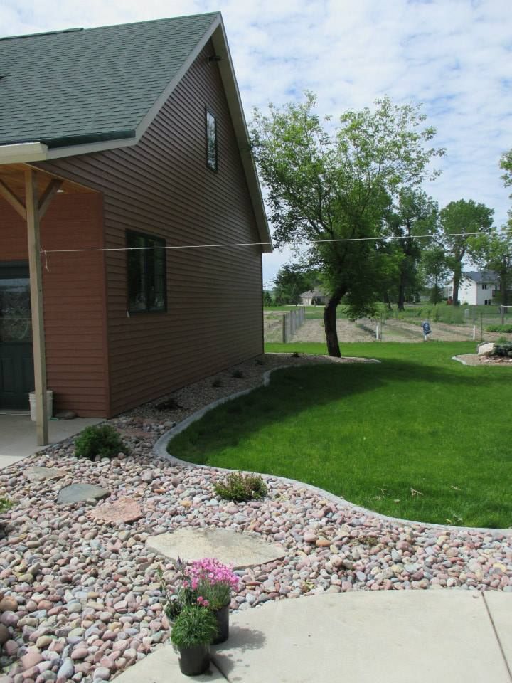 A house with brown siding and green roof, landscaped with rocks, grass, and a small tree.