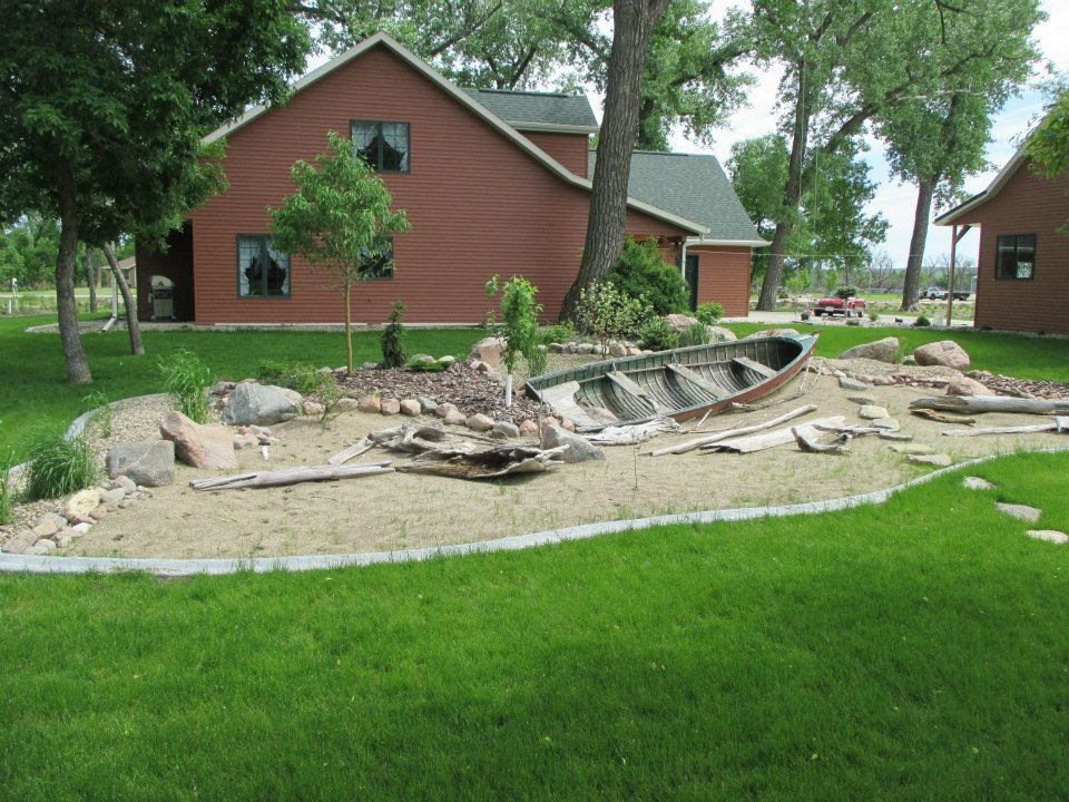 Brown house with a wooden boat in a sand garden on green grass.