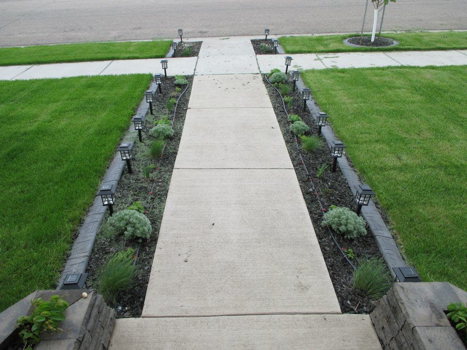 Concrete walkway leading to a house, flanked by flowerbeds with solar lights and green grass.
