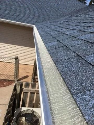 A silver gutter with a protective mesh cover runs along the edge of a shingled roof above an outdoor air conditioner.