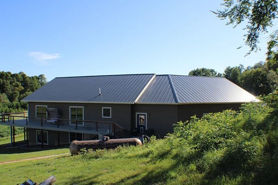 A dark-sided house with a metal roof and a raised deck, situated on a grassy, partially wooded lot under a clear blue sky.