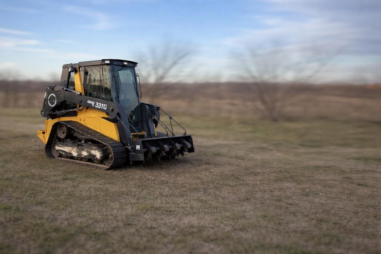 Yellow-and-black tracked skid steer on a grassy field with an overcast sky